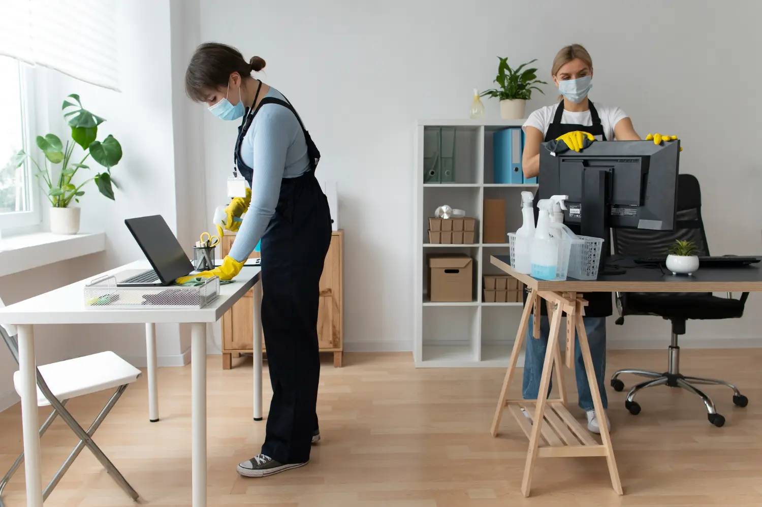 Two cleaners disinfect office desks and computer equipment wearing masks